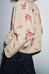 Chaqueta acolchada color beige con bordados florales en tonos rosados y rojos. Tiene escote en pico, manga larga y forro interior rojo. El cierre frontal es con alamar y tiene ribetes rojos.