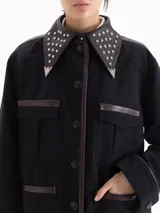 Chaqueta negra de corte recto con largo hasta la segunda cadera. Presenta detalles de cuero en cuello, frente, hombros y puños, con cuello decorado con tachas metálicas y punteras. Cuenta con bolsillos frontales tipo cargo.