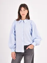 Camisa de manga larga en color celeste, con cuello clásico y cierre frontal de botones. Presenta un diseño con mangas abullonadas y puños ajustados.