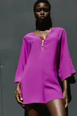 Mono corto de color fucsia, con escote en pico delantero y en la espalda, mangas anchas y largas, y detalle de fornitura dorada en el escote delantero con tiras finas para atar en la espalda.