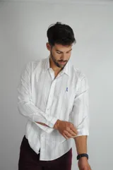 Camisa de manga larga con diseño de rayas verticales finas en tono azul sobre fondo blanco. Presenta cuello clásico, cierre frontal con botones y logo bordado en el pecho.