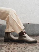 Bota corta de cuero color gris y beige, con detalle de correa marrón con hebilla y tachas plateadas. Tiene suela de goma negra y taco de madera.