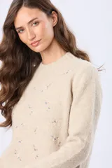 Buzo tejido de punto en color beige, con cuello redondo, mangas largas y puños acanalados. Presenta un delicado diseño bordado con motivos florales dispersos en el frente y una etiqueta de cuero sintético en el ruedo.