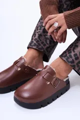 Zueco de cuero sintético color marrón con interior forrado en corderito sintético. Presenta una tira con hebilla metálica ajustable en el empeine y una suela de plataforma dentada en color negro.