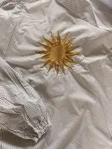 Camisa de corte suelto con estampado de rayas finas, cuello redondo y mangas largas con puños elásticos. Presenta un bordado central en la espalda con diseño de sol en color amarillo.