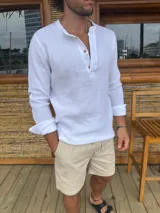Camisa de muselina blanca con cuello mao y cierre frontal parcial de botones. Presenta mangas largas con detalle de trabilla para arremangar y un corte ajustado al cuerpo.