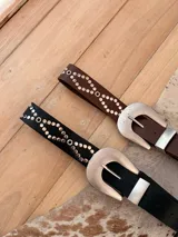 Cinturon de cuerina negra con tachas plateadas dispuestas en patron geometrico y hebilla metalica grande de estilo western.