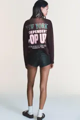 Camiseta de manga larga color marrón oscuro con efecto lavado. Presenta un cuello redondo y un estampado en el pecho con el texto "NEW YORK" en verde y "POP UP" en rosado.