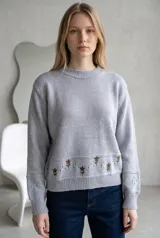 Sweater de punto color crema con cuello redondo, manga larga y diseño de calados en los puños. Presenta un detalle de bordado floral en la parte inferior y una franja con perforaciones decorativas.