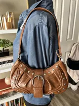 Bolso de hombro negro con textura de cuero y tachuelas plateadas. Presenta un asa corta y una hebilla plateada en la parte delantera.