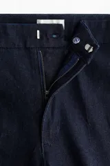 Jean de tiro alto con corte acampanado desde la rodilla. Presenta un diseño de denim oscuro con pliegues frontales y traseros, cierre con botón oculto y bolsillos traseros decorativos.