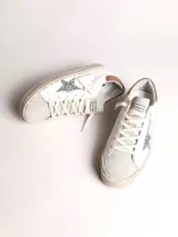 Zapatillas urbanas Golden Goose modelo Hi Star, de cuero blanco con detalles en gris y marrón. Presentan una estrella plateada con glitter en el lateral y suela de goma con inscripciones.