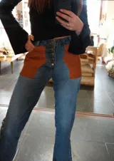 Pantalón de jean celeste con cintura elastizada, corte recto, bolsillos laterales y aplique de cuero marrón en el frente. Presenta botones metálicos en los laterales y etiqueta de cuero en la parte posterior.