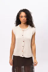 Chaleco de punto color beige, sin mangas, con cuello redondo y cierre frontal con botones de madera. Presenta un tejido de punto liso y terminaciones acanaladas en cuello y sisas.