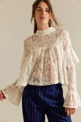 Top de encaje calado blanco, de corte corto y diseño amplio, con cuello alto, canesú frontal, volados superpuestos en el pecho y mangas largas acampanadas con detalle de volados.