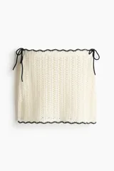 Pollera mini playera de punto crochet color blanco, de tiro bajo con tiras finas ajustables en los laterales y ribetes festoneados negros en contraste.