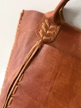 Cartera tipo shopper de cuero genuino marrón, con terminaciones artesanales trenzadas en las asas y los laterales, bolsillo frontal externo y asas largas para llevar al hombro.