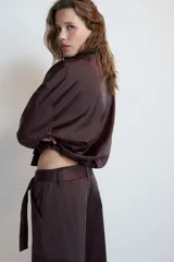 Cazadora satinada de color negro, con cuello subido, manga larga y bolsillos frontales de plastrón con cordones decorativos. Cuenta con bajo elástico y cierre frontal con cremallera y botones a presión oculta.