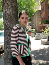Chaqueta corta tejida a rayas horizontales marrones y blancas, con cuello redondo y ribete verde. Tiene bolsillos frontales y mangas 3/4.