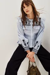 Camisa de algodón celeste con detalles de volados en los hombros y bordados étnicos en color negro en el pecho y las mangas. Presenta cierre frontal con botones y puños ajustados.