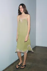 Vestido midi de satén color verde oliva, con escote recto y tirantes finos ajustables. Presenta detalles de encaje en color crema en el escote y en el bajo, con aberturas laterales.