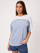 Remera oversize de manga corta marca Rusty, modelo Rocca, en combinación de colores celeste, blanco y detalles en verde. Presenta estampado frontal con el logo de la marca y diseño de bloques de color.