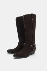 Bota alta de serraje marrón oscuro, de estilo texano, con tacón en bloque y puntera ligeramente puntiaguda. Cuenta con detalle de tiras cruzadas con tachas metálicas y una arandela en el tobillo, además de trabillas laterales en la parte superior de la caña.