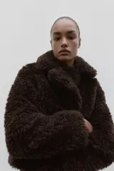 Abrigo corto de textura de pelo sintético, color marrón oscuro, con cuello de solapa, manga larga y cierre frontal con corchetes.