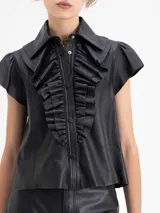 Camisa de cuero ovino color gris claro, con cuello camisero, volados centrales y mangas cortas con volumen generado por frunces en la copa. Cierre frontal con broches forrados.