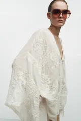 Blusa tipo capa de algodón con escote en pico y cierre frontal con botones. Presenta mangas largas amplias y diseño con bordados florales a tono, con bajo asimétrico.