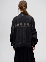 Campera tipo bomber confeccionada en denim de algodón color negro. Presenta un diseño holgado con cierre frontal metálico, bolsillos laterales con cierre y detalles decorativos en forma de escudos en la parte superior delantera.