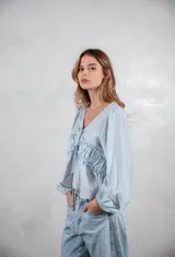 Camisa de tela liviana color celeste, con escote en V y cierre frontal mediante lazos ajustables. Presenta un diseño con volados horizontales en el pecho y mangas largas abullonadas con puños elásticos.
