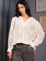 Blusa de tela liviana color crudo, con bordados florales tono sobre tono, escote en V con detalle de puntilla central, mangas largas con volumen y puños camiseros.