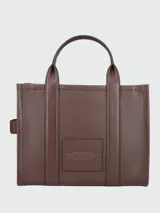 Cartera tipo tote de cuero granulado color tostado, con diseño estructurado y rectangular. Presenta doble asa superior fija y una correa larga ajustable y extraíble. En el frente, destaca el logo estampado con la inscripción 'THE TOTE BAG MARC JACOBS'.