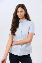 Camisa celeste rayada de manga corta, con cuello camisero y cierre frontal con botones. Presenta un corte entallado y un pequeño detalle de placa metálica en el borde inferior.