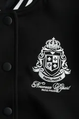 Campera de béisbol negra con cuello, puños y bajo acanalados con rayas blancas. Presenta un escudo bordado en blanco en el pecho.