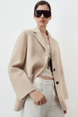 Abrigo corto de Zara confeccionado en hilatura con lana, de color beige. Presenta cuello solapa, manga larga, cierre frontal con botones negros y bolsillos de plastrón en el delantero.