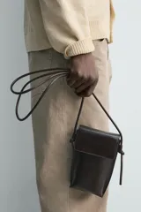 Bandolera porta teléfono de piel marrón con acabado liso, compartimento principal con solapa imantada y asa de hombro ajustable con nudos.