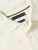 Chomba color crema con patrón integral de logos de la marca. Tiene cuello tipo polo, cartera de dos botones, mangas cortas y logo de Tommy Hilfiger bordado en la manga. Confeccionada en algodón BCI y elastolefín.