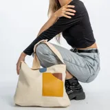 Bolso bandolera de lona color beige, con correa ajustable y diseño de parches en tonos amarillo y marrón en el frente. Cuenta con un detalle de etiqueta azul en la parte superior.
