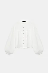 Blusa corta de color blanco con diseño de bordados perforados, cuello solapa, cierre frontal con botones y mangas largas abullonadas.