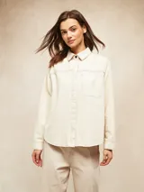 Camisa de manga larga color crudo, con cuello clásico, botones frontales y bolsillo de parche en el pecho. Presenta costuras decorativas en contraste.