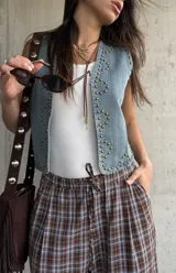 Chaleco de jean color celeste claro, con escote en V, sin mangas y decorado con tachas metálicas doradas distribuidas en los bordes y formando diseños en los paneles frontales.