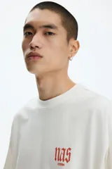 Remera de algodón blanco crudo, de corte oversized con hombros caídos y cuello redondo acanalado. Presenta pequeño estampado en el pecho con la inscripción "nas ©1994" en tono anaranjado.