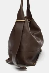 Bolso tipo saco de gran tamaño en cuero marrón oscuro, con textura granulada. Presenta un asa de hombro ancha y detalles metálicos dorados en la unión del asa. Incluye un bolsillo interior extraíble y cierre mediante botón imantado.
