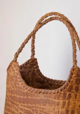 Bolso tipo matera de cuero con textura de reptil, color tostado, con manijas trenzadas y detalles de costura artesanal en los bordes.