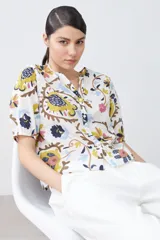 Camisa blanca con estampado floral en tonos azul, amarillo, rosa y marrón. Tiene cuello mao, mangas cortas abullonadas y cierre frontal con botones.