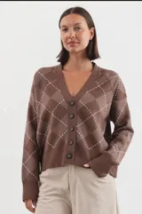 Cardigan de punto con escote en V y cierre frontal de botones. Presenta un diseño de estampado argyle en tonos marrones con líneas blancas. Posee mangas largas con puños acanalados y ruedo también acanalado.