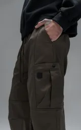 Pantalón cargo de corte regular confeccionado en textil técnico pesado color chocolate. Presenta cintura elástica con cordón de ajuste, dos bolsillos laterales en la cintura y dos bolsillos cargo de gran tamaño. Incluye elásticos con tancas en los tobillos para regular la apertura de la pierna.