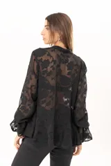 Blusa negra de tejido semitransparente con diseño floral en relieve, cuello alto con lazo frontal y mangas largas con volados en los puños.